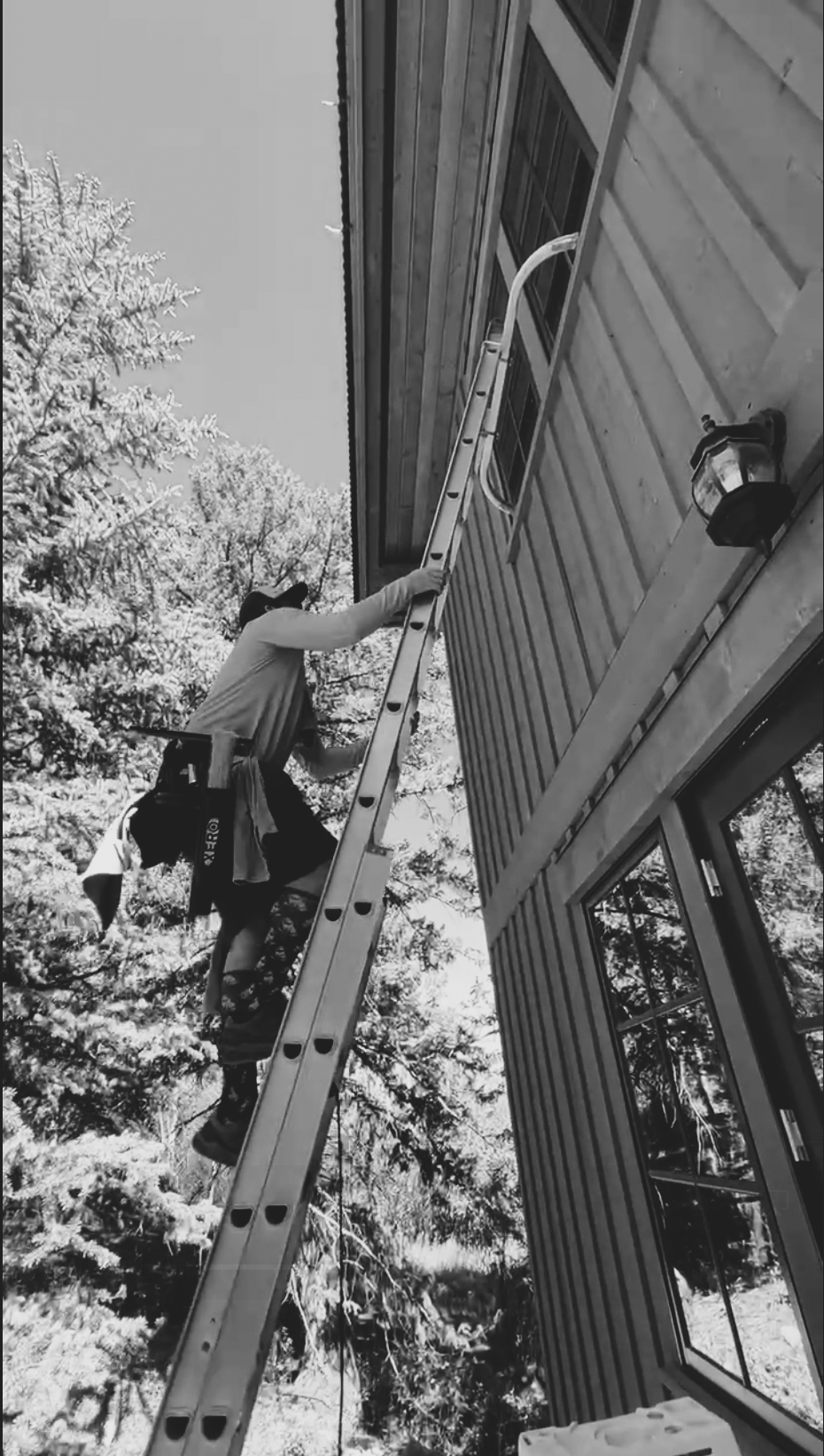 Black-and-white photo of a window cleaner climbing a tall extension ladder to reach upper-story windows on a wood-sided mountain home at Arrowhead in Edwards, Colorado